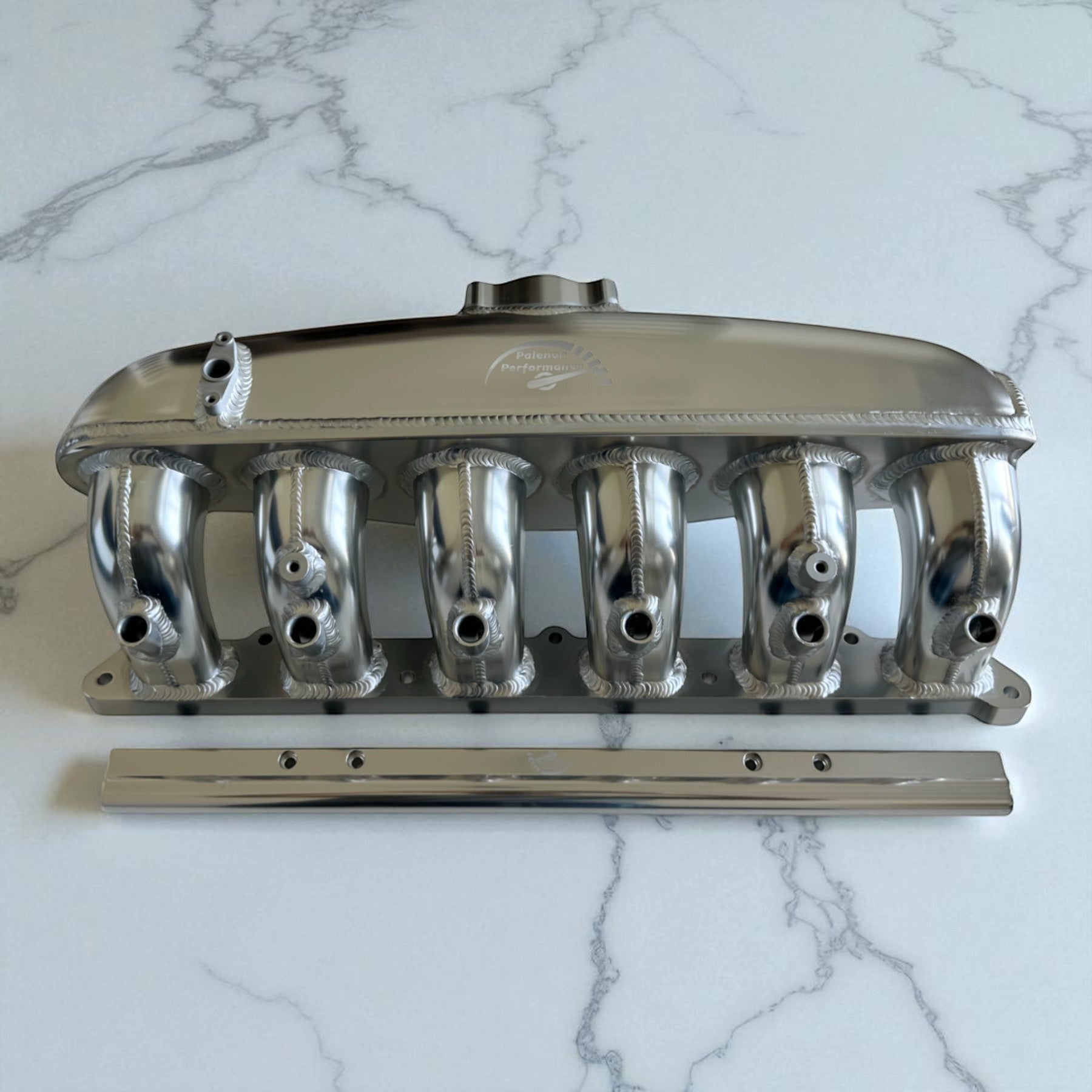 Palenon Performance aluminum N54 intake manifold with port injection bungs for BMW engines on a marble surface.